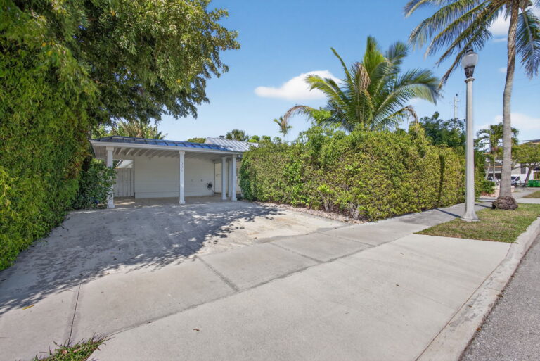 Concrete driveway leading to white carport, bordered by tall privacy hedges, with sidewalk and street lamp in foreground
