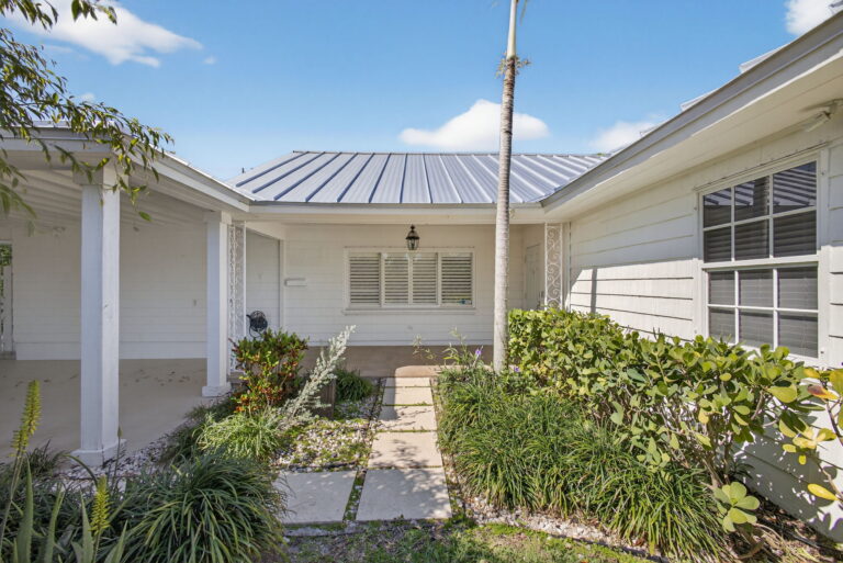 Front exterior with white siding, grey metal roof, stone walkway, tropical landscaping, and palm tree under a blue sky.