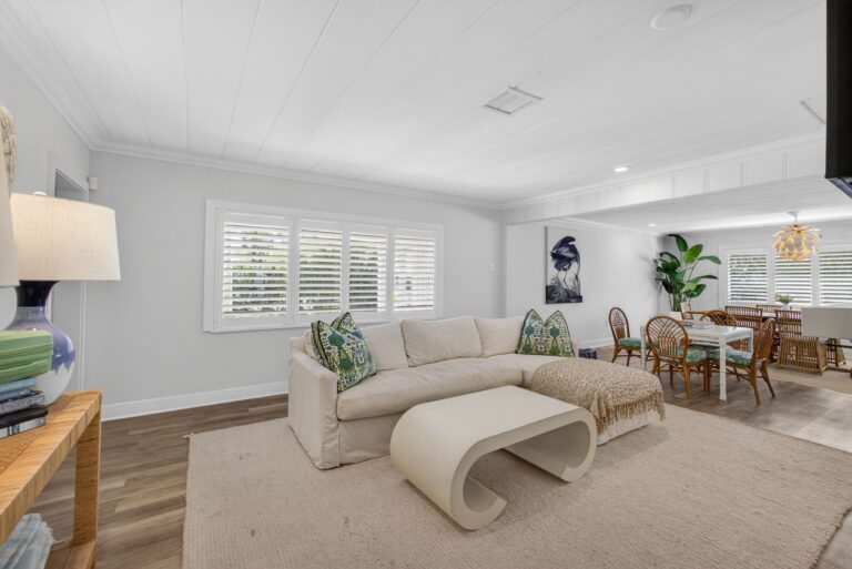 Open-concept living room with white sectional sofa, curved white coffee table, wood-look floors, plantation shutters, and dining area in background.