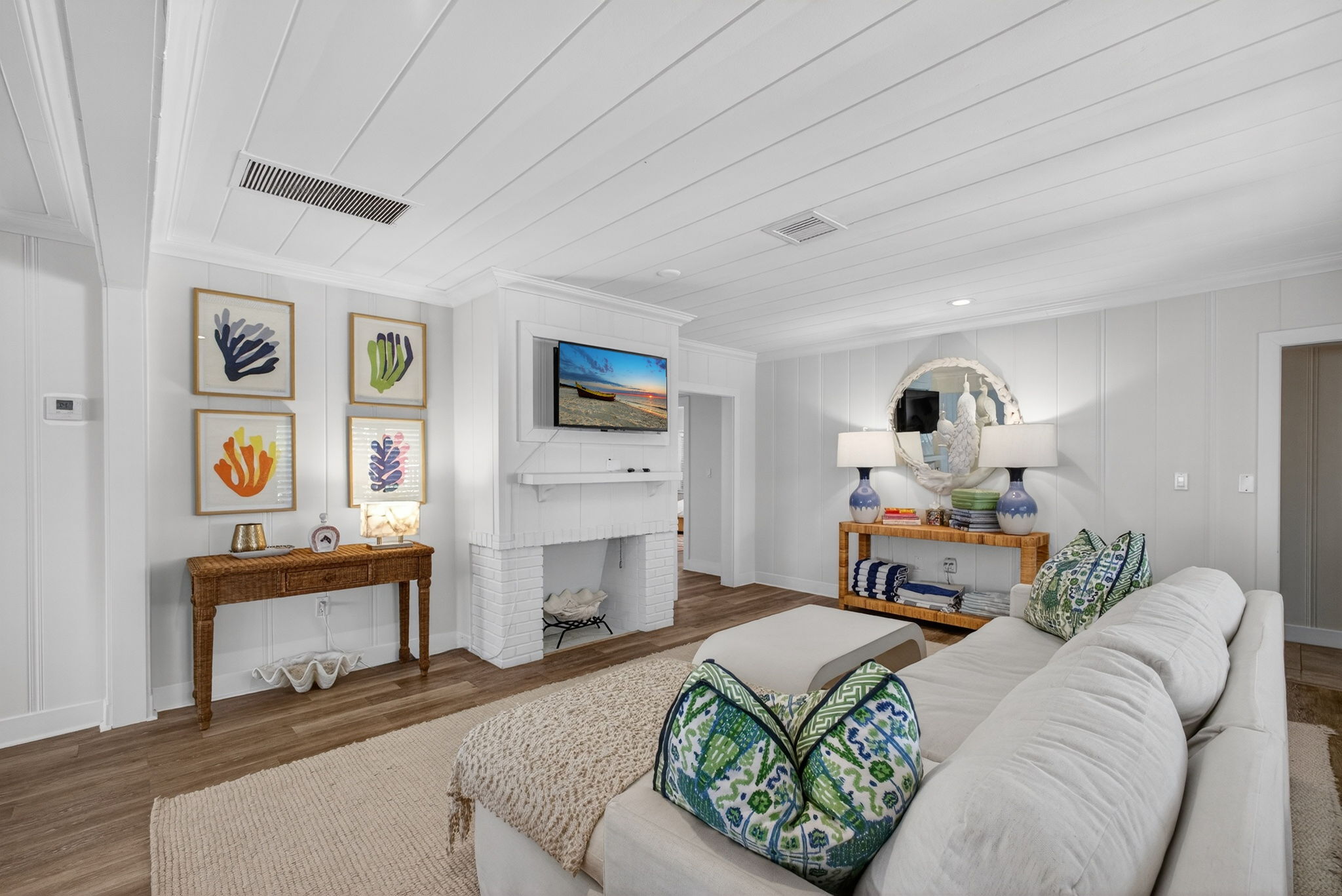 Bright white living room with sectional sofa, white fireplace, wall-mounted TV, light wood floors, and colorful botanical wall art
