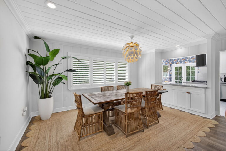 Dining room with rustic wood table, wicker chairs, round jute rug, gold artichoke pendant light, potted plant, and window with plantation shutters.