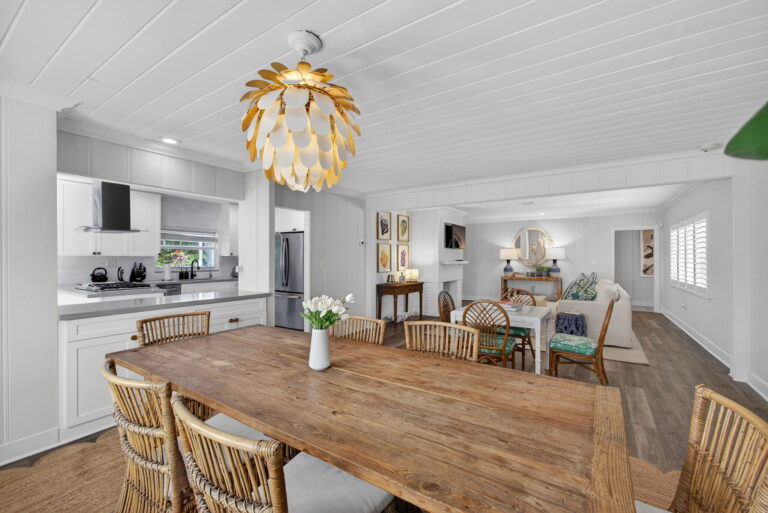 Dining area with rustic wood table, wicker chairs, gold leaf pendant light, and open view to white kitchen and living room.