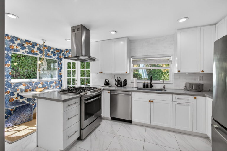 Modern white kitchen with grey countertops, stainless steel range and hood, gold knobs, and blue floral accent wallpaper.
