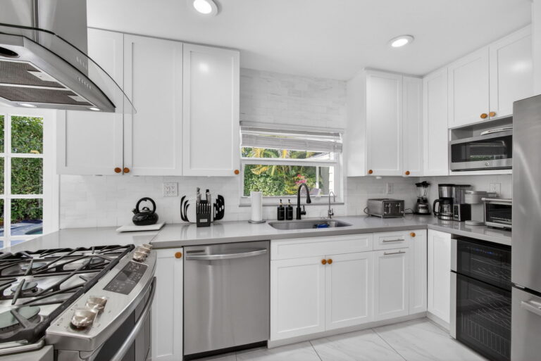 Modern white kitchen with shaker cabinets, light countertops, stainless steel appliances, white tile backsplash, and garden view through the sink window