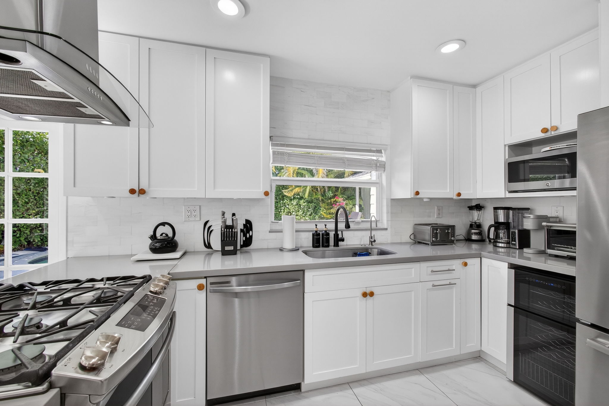 Modern white kitchen with shaker cabinets, light grey countertops, stainless steel appliances, and garden view through the sink window