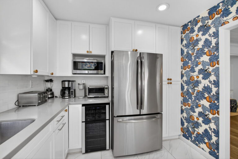 Kitchen detail with stainless steel French-door refrigerator, built-in microwave, wine cooler, white cabinets, and floral wallpaper accent wall.