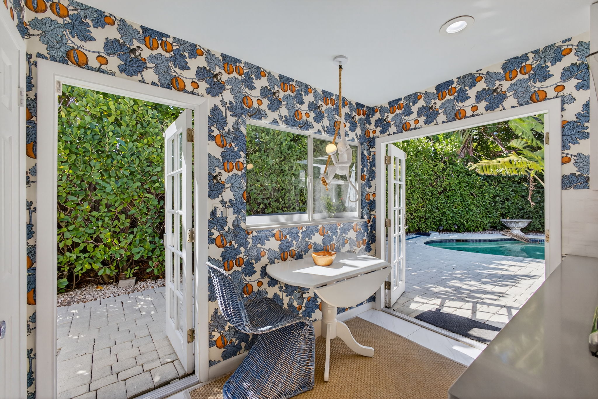 Sunlit dining nook with blue and orange floral wallpaper, white round table, blue wicker chairs, and open French doors to the patio and pool.