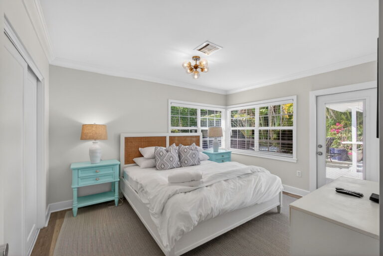 Bright bedroom with white bedding, wood-and-wicker headboard, light blue nightstands, plantation shutters, and glass door to the outdoors.