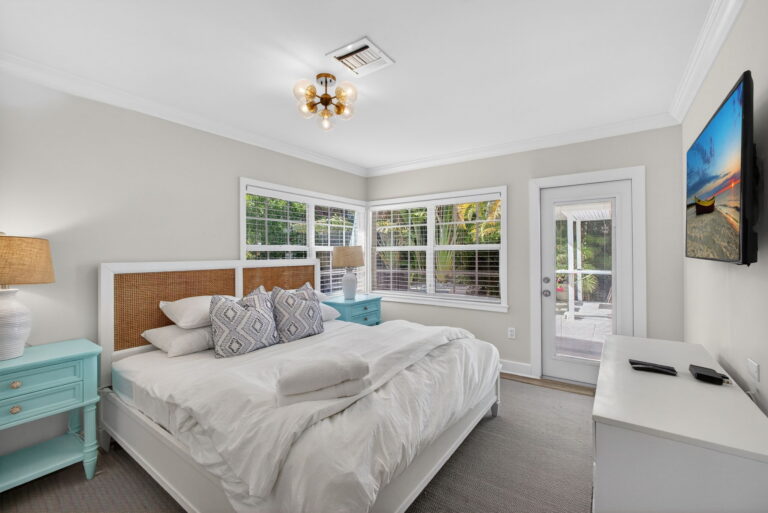 Primary bedroom with white bedding, wood-and-wicker headboard, light blue nightstands, windows with tropical view, glass door, and wall-mounted TV above white dresser.