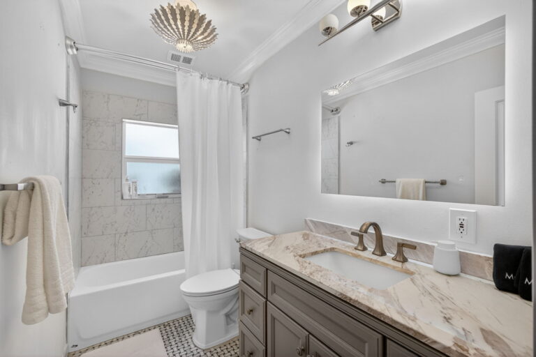 Bathroom with grey vanity, marble countertop, frameless mirror, three-bulb light fixture, tub-shower combo, and black-and-white hexagonal floor tile.