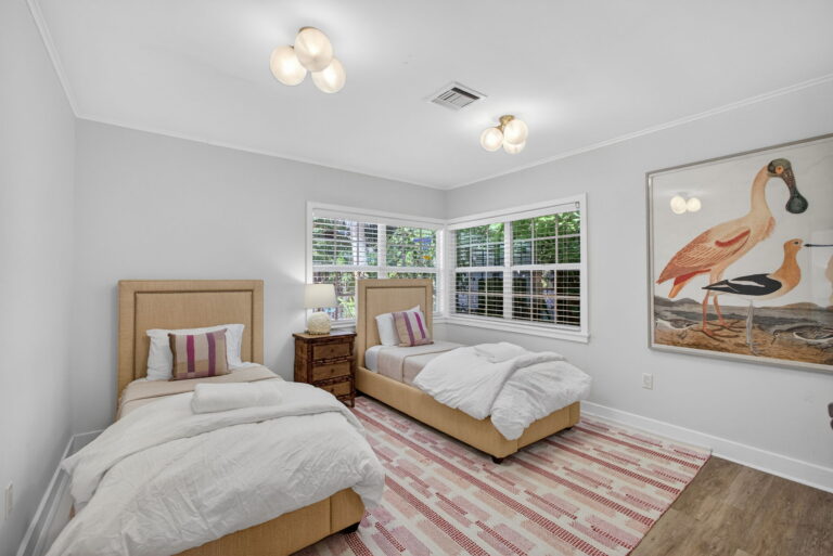 Wide twin bedroom view with flamingo art, pink-and-white rug, two twin beds, and bright windows with white shutters.