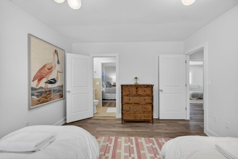 Bedroom view toward open doorways, large pink flamingo wall art, hallway with wood dresser, and patterned area rug.