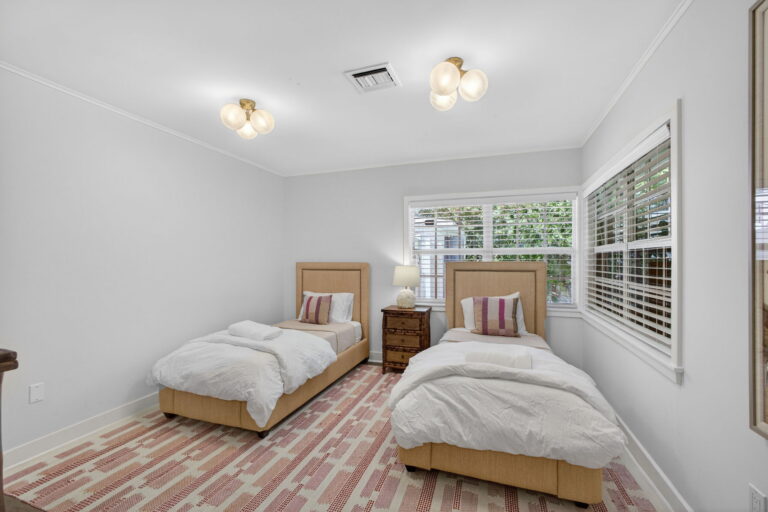 Twin bedroom with tan upholstered headboards, white bedding, dark wood nightstand, pink-and-white patterned rug, and plantation shutters.