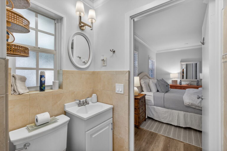 Bathroom with tan wall tile, white pedestal vanity, toilet, round mirror, and open doorway to bedroom with white bedding.