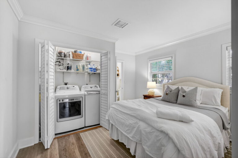 Bedroom with white bedding, open bifold closet doors revealing stacked washer and dryer, and shelving in compact laundry nook.