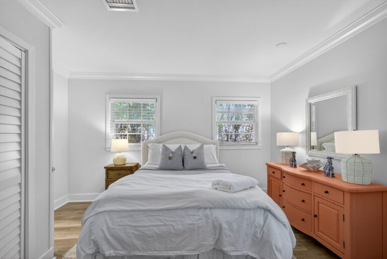 Second bedroom with white arched headboard, blue-striped pillows, coral nine-drawer dresser, white lamps, and windows with white blinds.