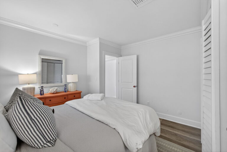 Wide view of second bedroom from foot of bed, showing coral dresser, striped accent pillow, and open doorway from hallway.