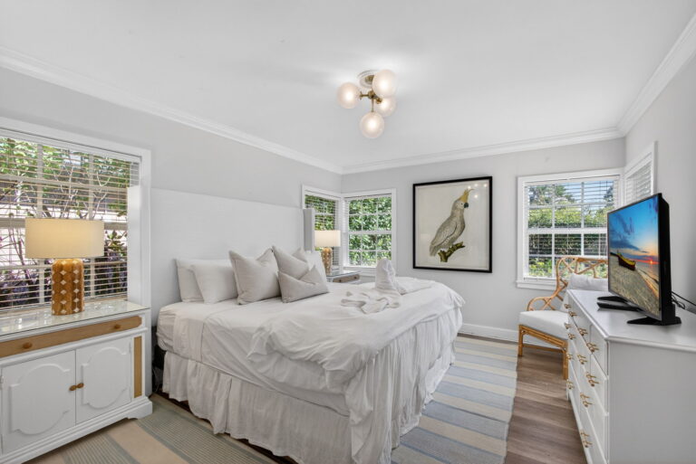 Bright bedroom with white bedding, gold lamp, white dresser with TV, cockatoo wall art, striped rug, and brass ceiling light fixture.