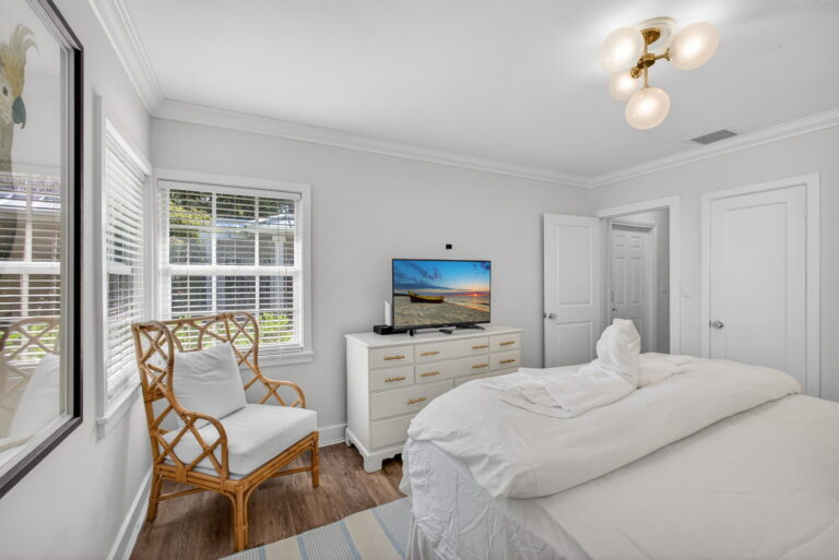 Bedroom corner with wicker armchair, window with white blinds, white dresser with TV, and open doorway to bathroom.