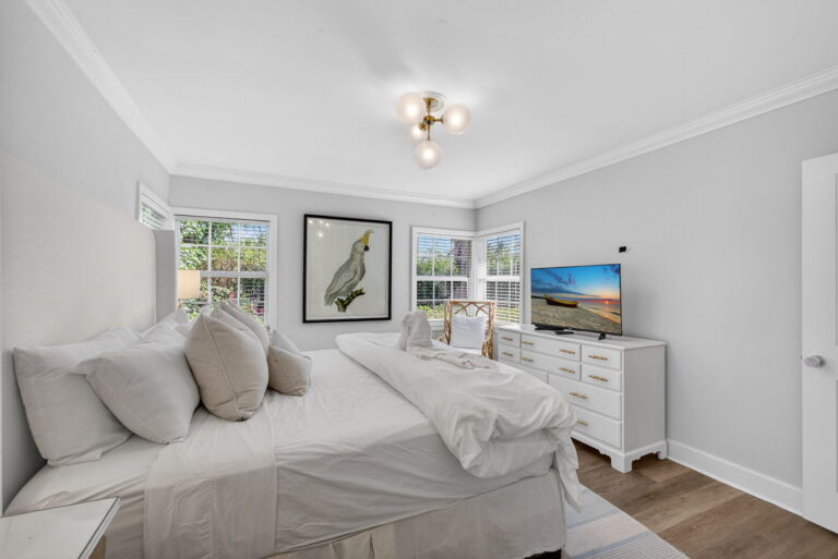 Side view of bedroom showing white headboard, cockatoo artwork, dresser with TV, and large window with natural light.