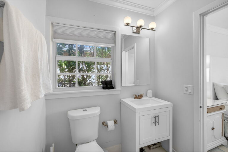 Clean white bathroom with vanity, square sink, toilet, mirror with three-bulb light, window with blinds, and towel rack.