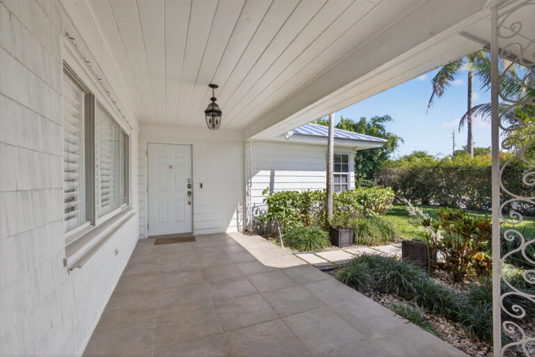 Covered white porch with tile flooring, black lantern light, white door, and view of landscaped garden and palm trees.