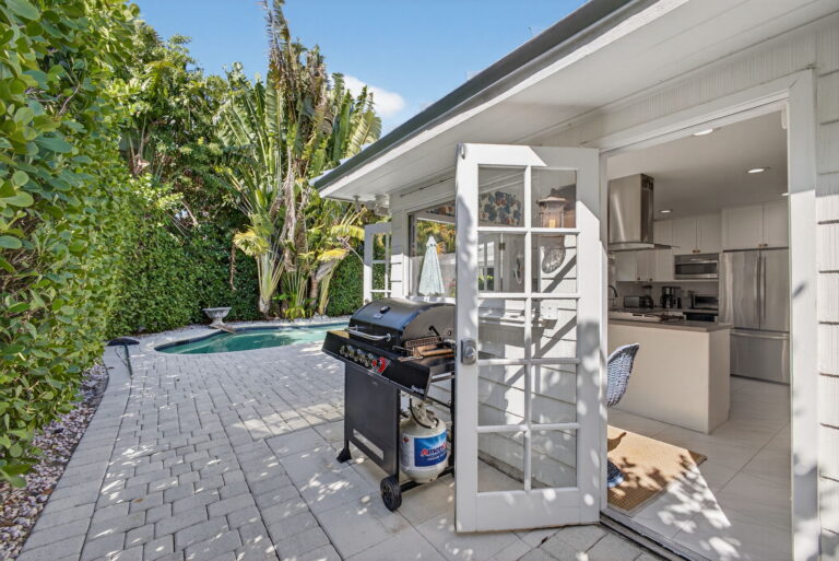 Stone patio with black gas grill, open French doors to white kitchen with island, pool edge, and tropical landscaping.