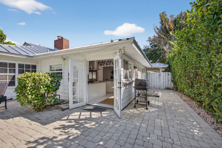 Wide backyard patio behind white home with metal roof, gas grill, privacy hedges, and light grey paved surface.
