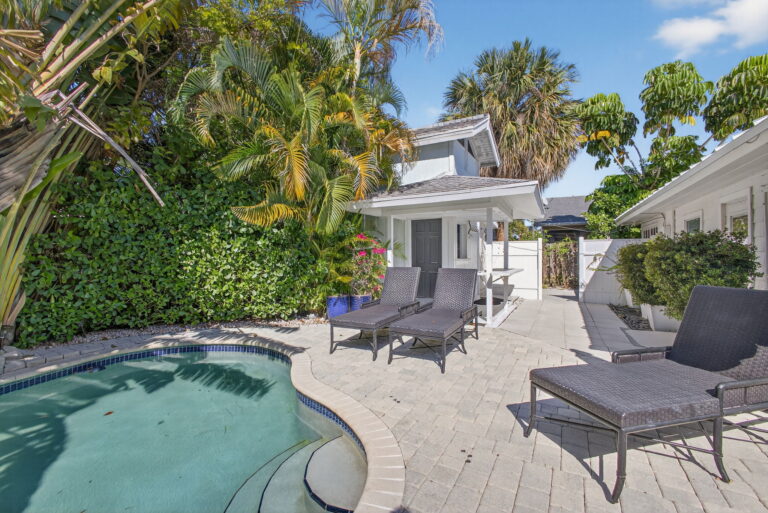 Wide backyard view with curved pool, blue tile, grey lounge chairs, outdoor bar, white home, and palm trees under blue sky.