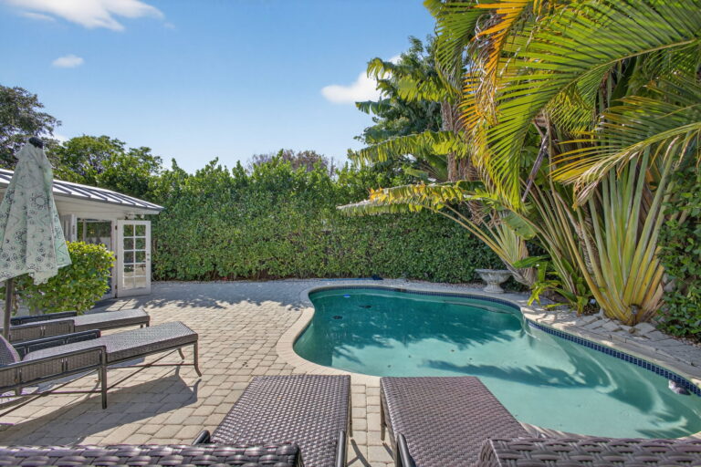 curved shaped pool with stone patio, wicker lounge chairs, white cottage with French doors, privacy hedge, and tropical palm landscaping.