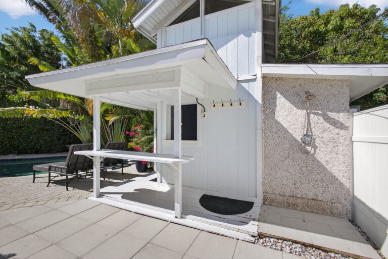 Outdoor bar with white frame and countertop, adjacent outdoor shower with textured wall, and grey lounge chairs near the pool.