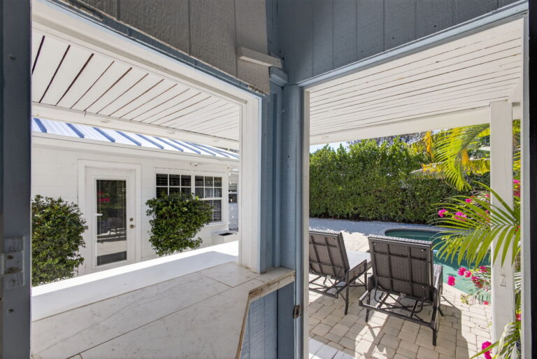 View from shaded covered structure across patio to pool, grey lounge chairs, white house, and tropical greenery.