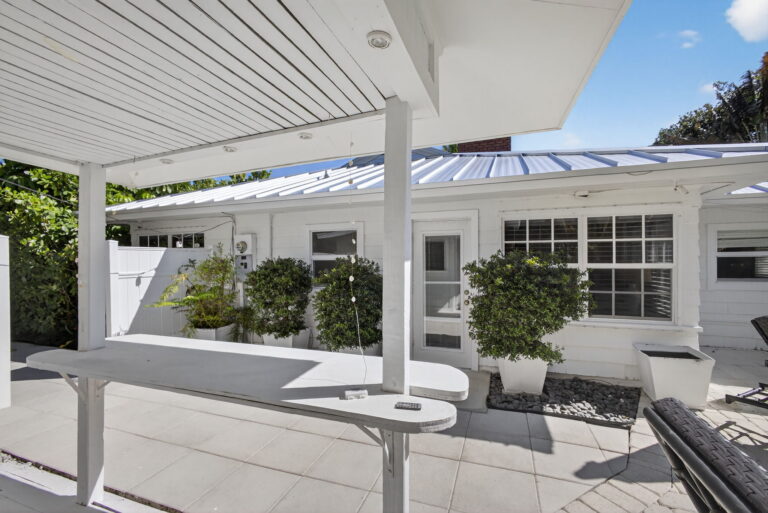 View from covered outdoor bar toward white house exterior, white door, windows, and three potted green shrubs lined along the wall.