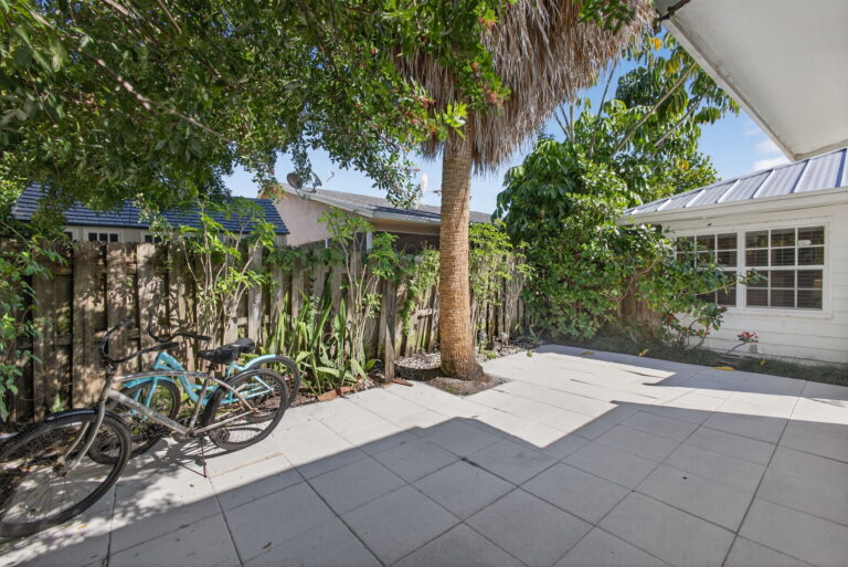 Shaded side patio with guest bicycles, palm tree, tiled flooring, wooden fence, and tropical plants along white exterior wall.