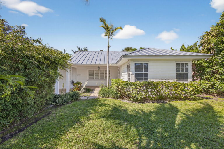 Front exterior of white single-story home with grey metal roof, green lawn, tropical landscaping, and palm trees under a clear blue sky.