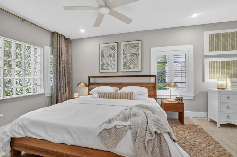 Primary bedroom with dark wood headboard, white bedding, botanical palm art, gold lamps, white dresser, and glass-block window.