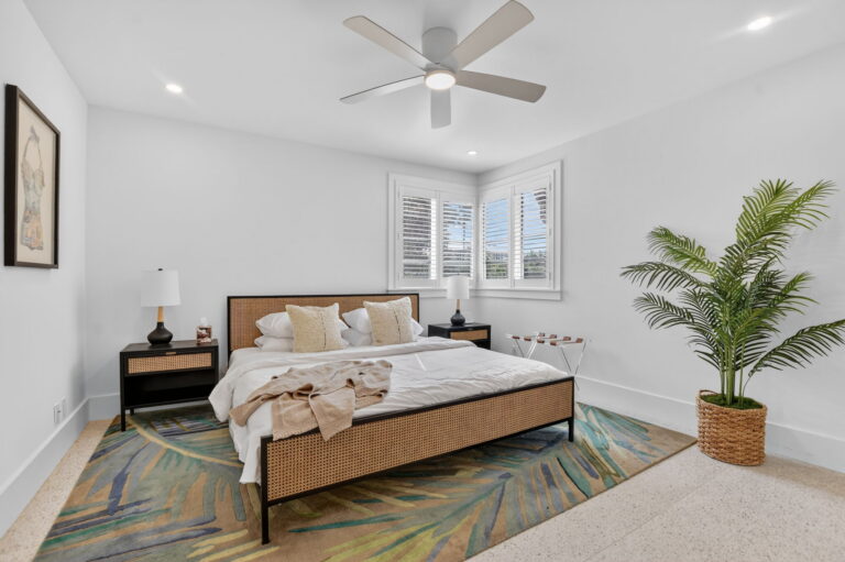 Wide bedroom view from the foot of the bed with tropical rug, palm plant in woven basket, cane headboard, and bright natural light.