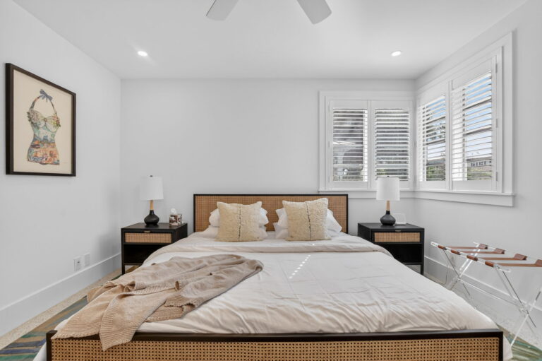Bedroom with woven cane bed frame, white bedding, black nightstands, plantation shutters, and vintage swimsuit wall art