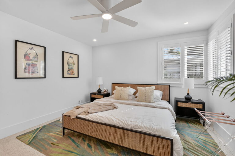 Angled bedroom view with tropical leaf rug, cane bed, ceiling fan, vintage swimsuit art, and luggage rack by the window.