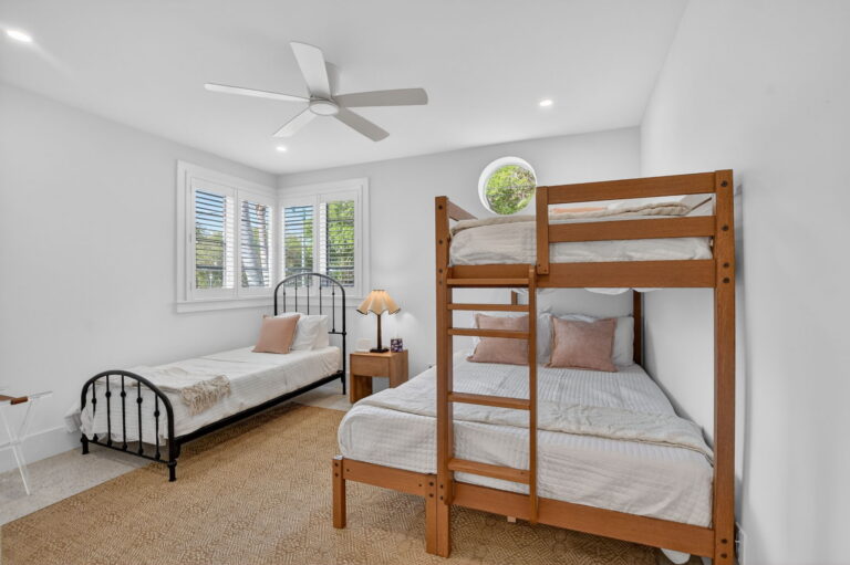 Guest bedroom with black metal twin bed, wooden bunk bed, white bedding, tan rug, plantation shutters, and ceiling fan.