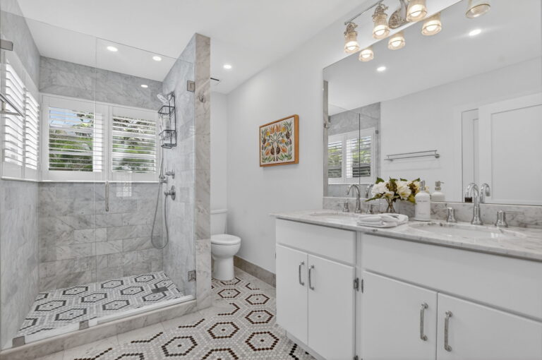 Modern bathroom with glass walk-in shower, marble tile walls, double vanity with grey marble countertop, hexagonal floor pattern, and white toilet.