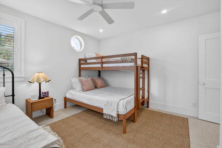 Angled view of the bunk bed bedroom with wooden ladder, nightstand, lamp, circular window, and woven tan area rug.