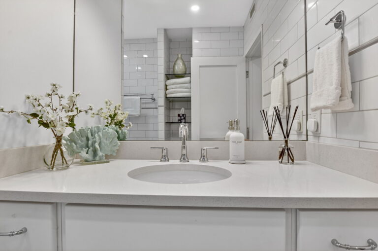 Close-up of bathroom vanity with white countertop, chrome fixtures, coral decor, subway tile wall, and towel niche reflected in the mirror.