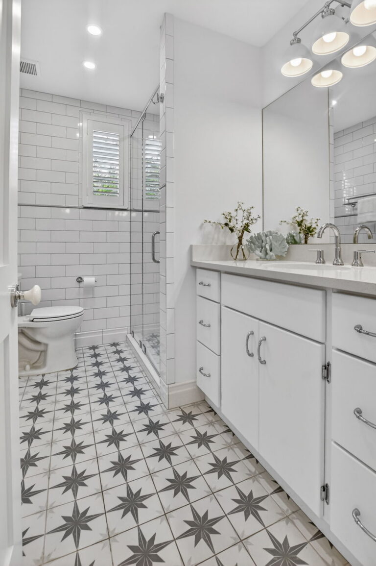 Full bathroom view with white toilet, plantation-shutter window, glass walk-in shower, subway tile walls, and patterned floor tile.