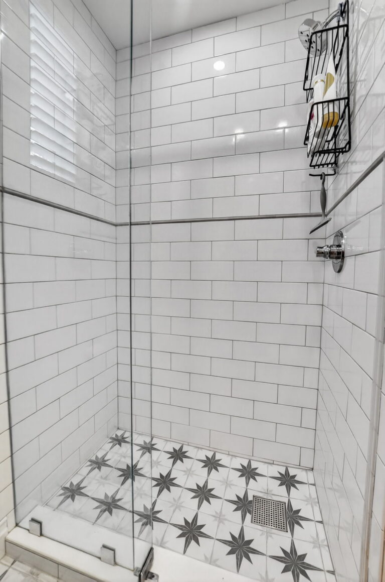 Walk-in shower with white subway tile, grey accent band, starburst floor pattern, chrome showerhead, and window with white blinds.
