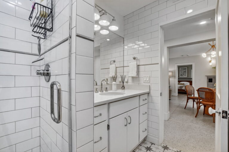 White subway-tile bathroom with vanity, patterned floor tile, walk-in shower, and view through the doorway to the dining area.