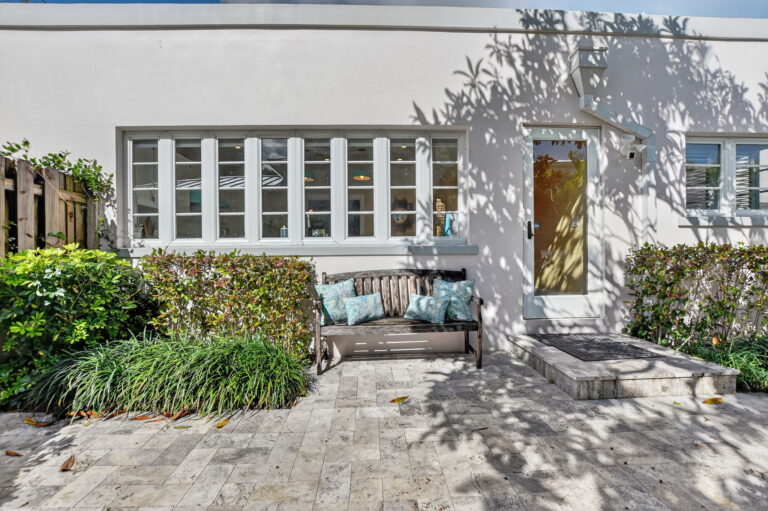 Back patio of the rental home with stone flooring, a wooden bench with blue cushions, glass door, and greenery along the white exterior wall.