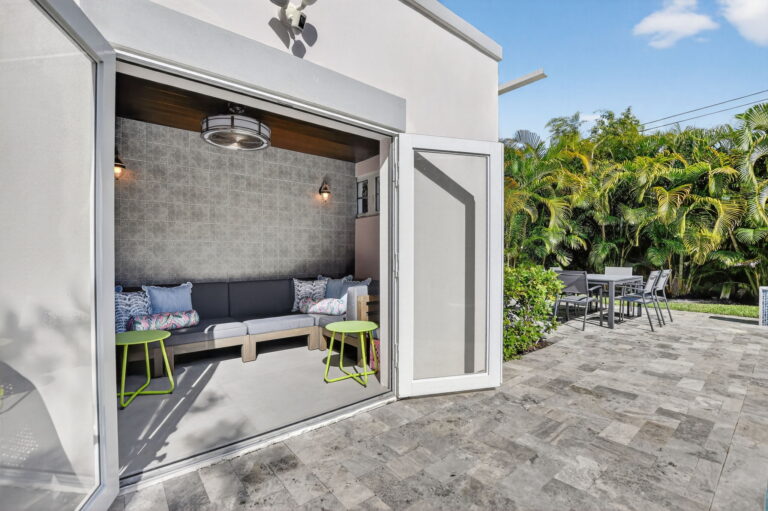 Open French doors from the living room to a covered patio with grey sectional sofa, lime-green side tables, and a sunny tropical backyard