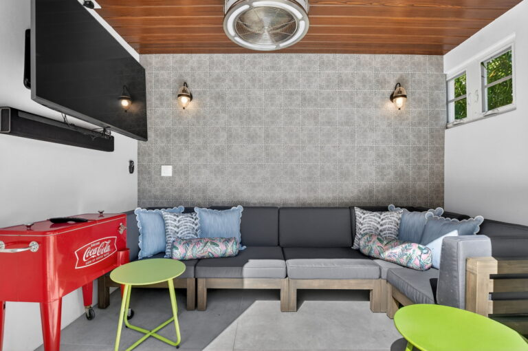 Covered outdoor lounge with grey sectional sofa, wall-mounted TV, tiled accent wall, wood ceiling, and a vintage-style red cooler