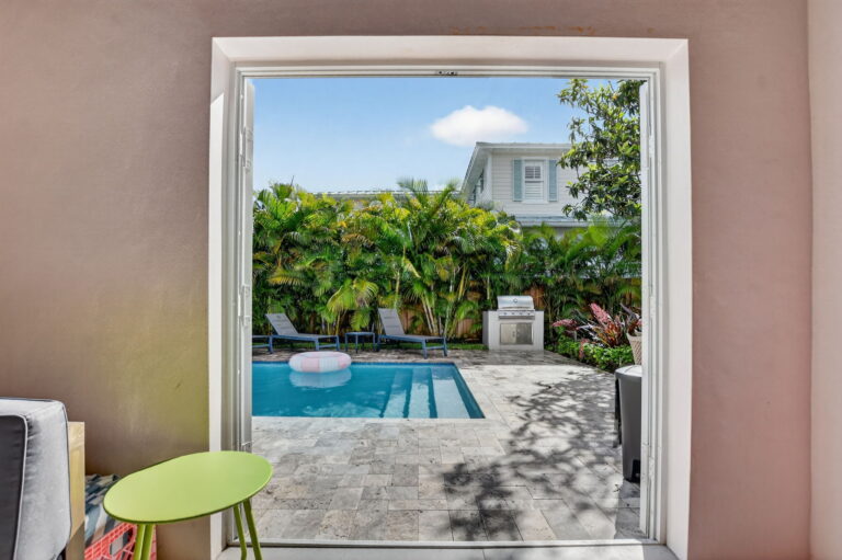 View from inside the home through open doors to the backyard pool, patio, outdoor grill, and tropical landscaping.